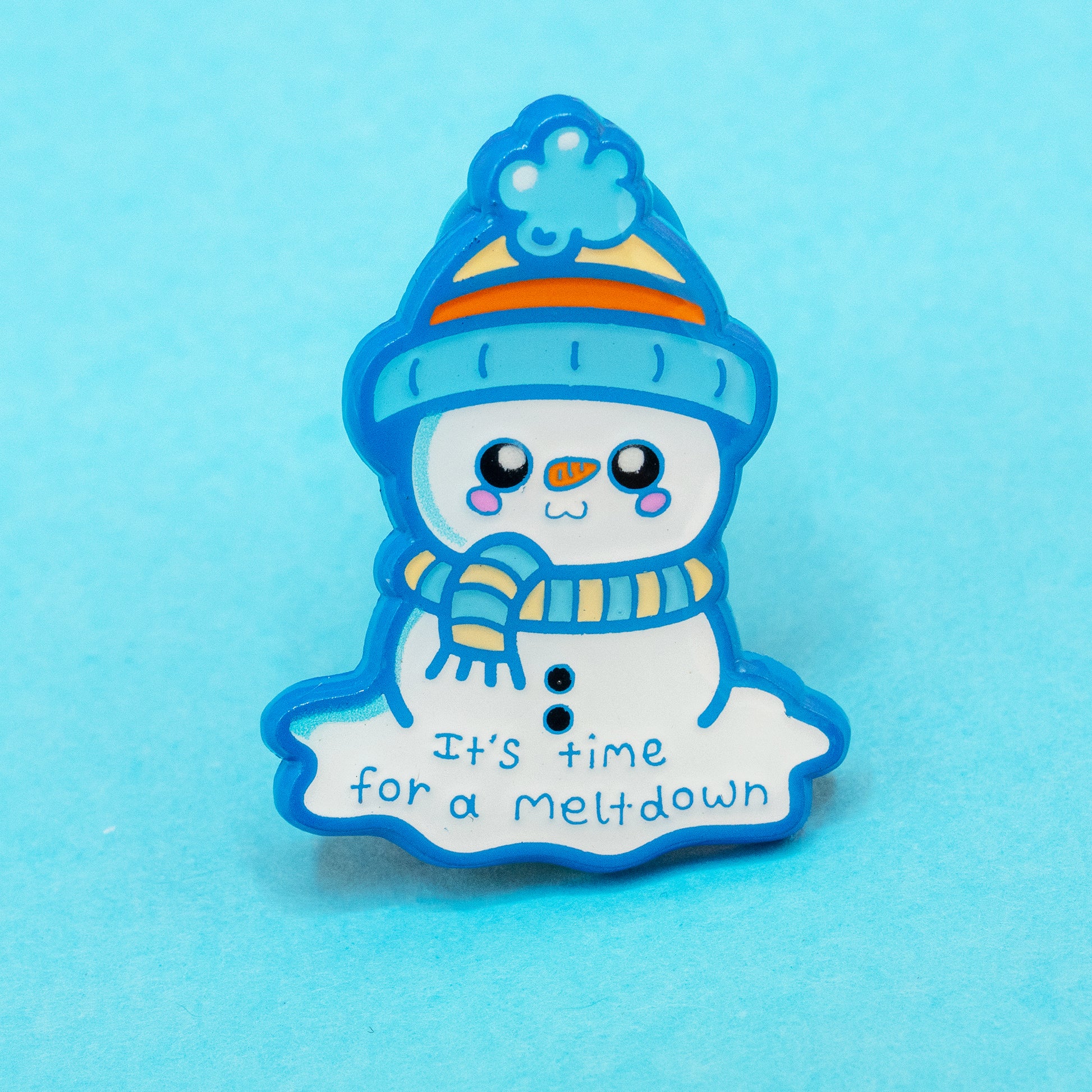 A close up of the Innabox ‘It’s Time For A Meltdown’ Snowman Enamel Pin against a bright blue background. The cute snowman design features a blue hat, scarf and a cheerful face with the phrase “It’s time for a meltdown” written beneath. A 30mm hard enamel pin designed to raise awareness for chronic illness, disability and mental health in a gentle and uplifting way.