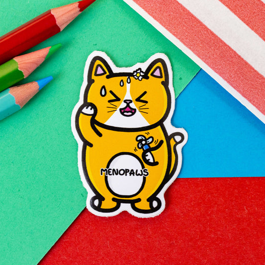 The Menopaws Cat Sticker - Menopause on a red, blue and green background with colouring pencils and red stripe candy bag. The orange cat shaped sticker is sweating and looks stressed holding a fan with a flower on its head. Across the middle is black text reading 'menopaws'. Raising awareness for menopause and menopausal symptoms.