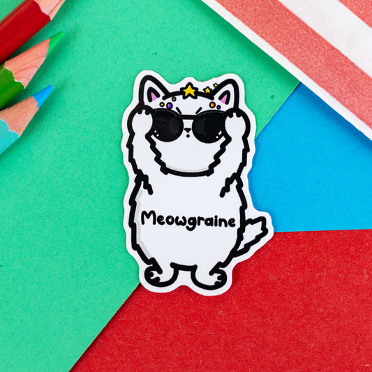 The Meowgraine Cat Sticker - Migraine on a red, blue and green background with colouring pencils and red stripe candy bag. A white stressed cat sticker clutching a pair of black sunglasses to its eyes with multicoloured spots and stars over its head, across its middle reads 'meowgraine'. The hand drawn design is raising awareness for migraines and headaches.