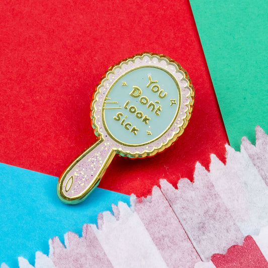 You Don't Look Sick Mirror Enamel Pin on a blue, teal and red background next to a red and white striped paper bag. The hand mirror shaped baby pink glittery enamel pin has gold text on it that reads 'you don't look sick'. The hand drawn design is made to raise awareness for invisible illnesses