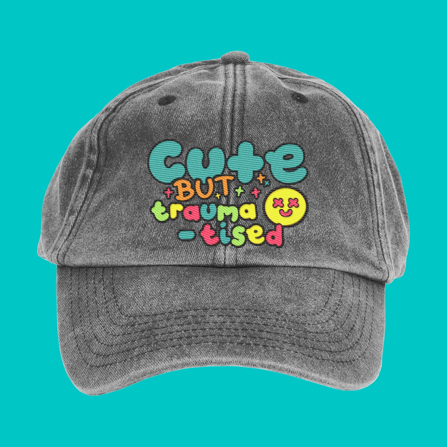 Front view of a vintage black-grey cap with colourful embroidered text reading "Cute but traumatised" in a playful font. The cap is unstructured with a curved peak, designed by Innabox to uplift those with chronic illness or hidden disabilities.