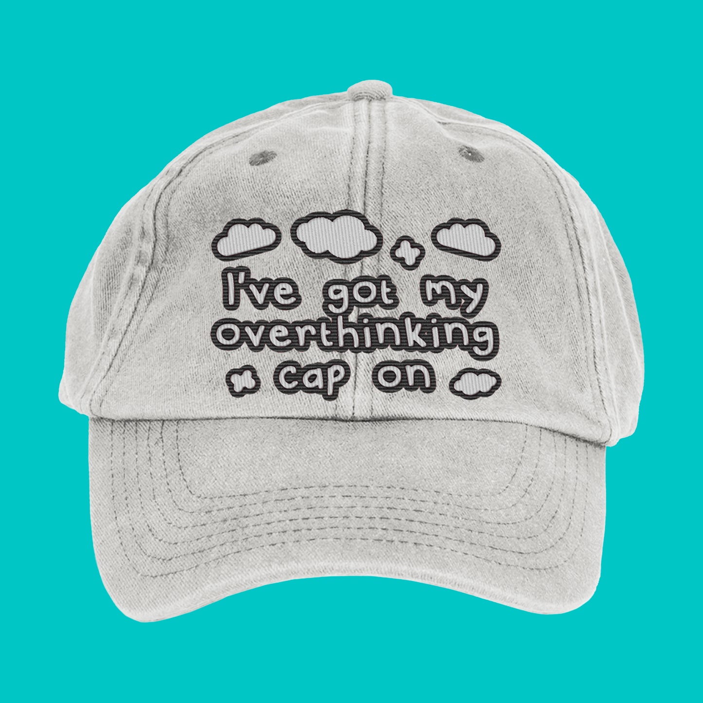 Close-up of the Innabox Overthinking Cap laid flat on a turquoise background. The vintage grey cap features black embroidered text and clouds that read “I’ve got my overthinking cap on.” A soft cotton 6-panel design with a curved peak and adjustable brass buckle strap.