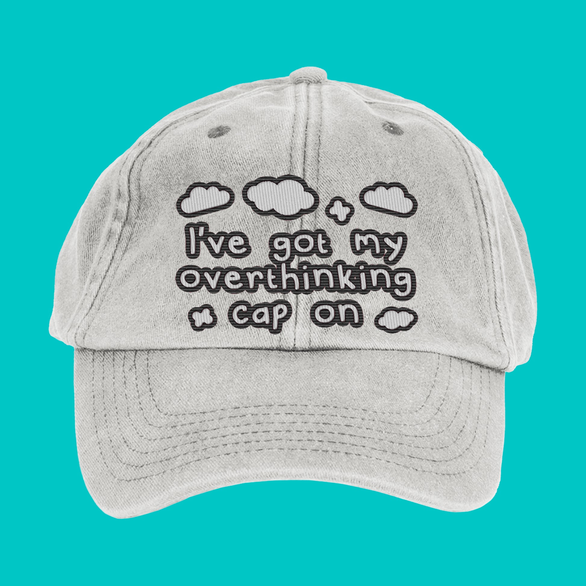 Close-up of the Innabox Overthinking Cap laid flat on a turquoise background. The vintage grey cap features black embroidered text and clouds that read “I’ve got my overthinking cap on.” A soft cotton 6-panel design with a curved peak and adjustable brass buckle strap.