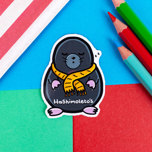 A sticker of a small grey mole looking sleepy wearing a yellow scarf it has text saying hashimoletos on its lower body. It is on a red, blue and green background with coloured pencils to one side and a stripy paper bag. The hand drawn design is raising awareness for Hashimotos.