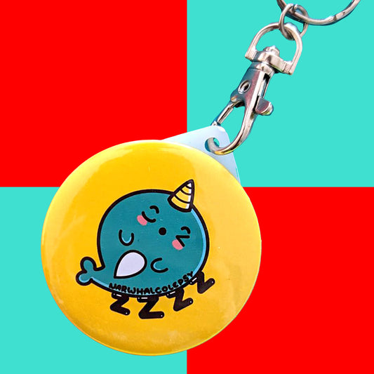 The Narwhalcolepsy Narwhal Keyring - Narcolepsy on a red and blue background. A silver lobster clip with a yellow plastic circular keychain of a sleeping blue narwhal on a bed of ZZZ with black text across its body reading 'narwhalcolepsy'. The hand drawn design is raising awareness for narcolepsy.