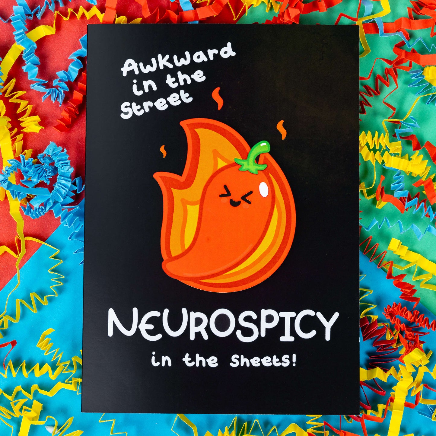 A black greeting card with a cute illustration of a red chilli pepper with scrunched up eyes and big smile. There is orange fire behind the chilli. 'Awkward in the street NEUROSPICY in the sheets!' is written on the card in white. The background of the photo is colourful card confetti. Design inspired by neurodivergent disorders.