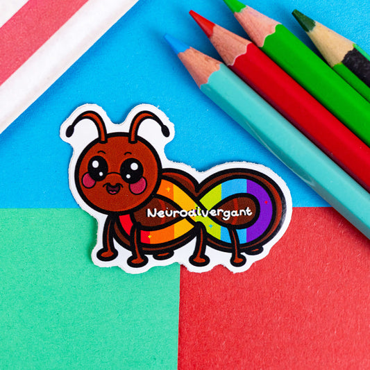 The Neurodivergant Ant Sticker - Neurodivergent on a red, blue and green background with colouring pencils and red stripe candy bag. The brown ant shape sticker is smiling with a rainbow infinity symbol and white text reading 'neurodivergant' with sparkles across its body. The hand drawn design is raising awareness for neurodiversity.