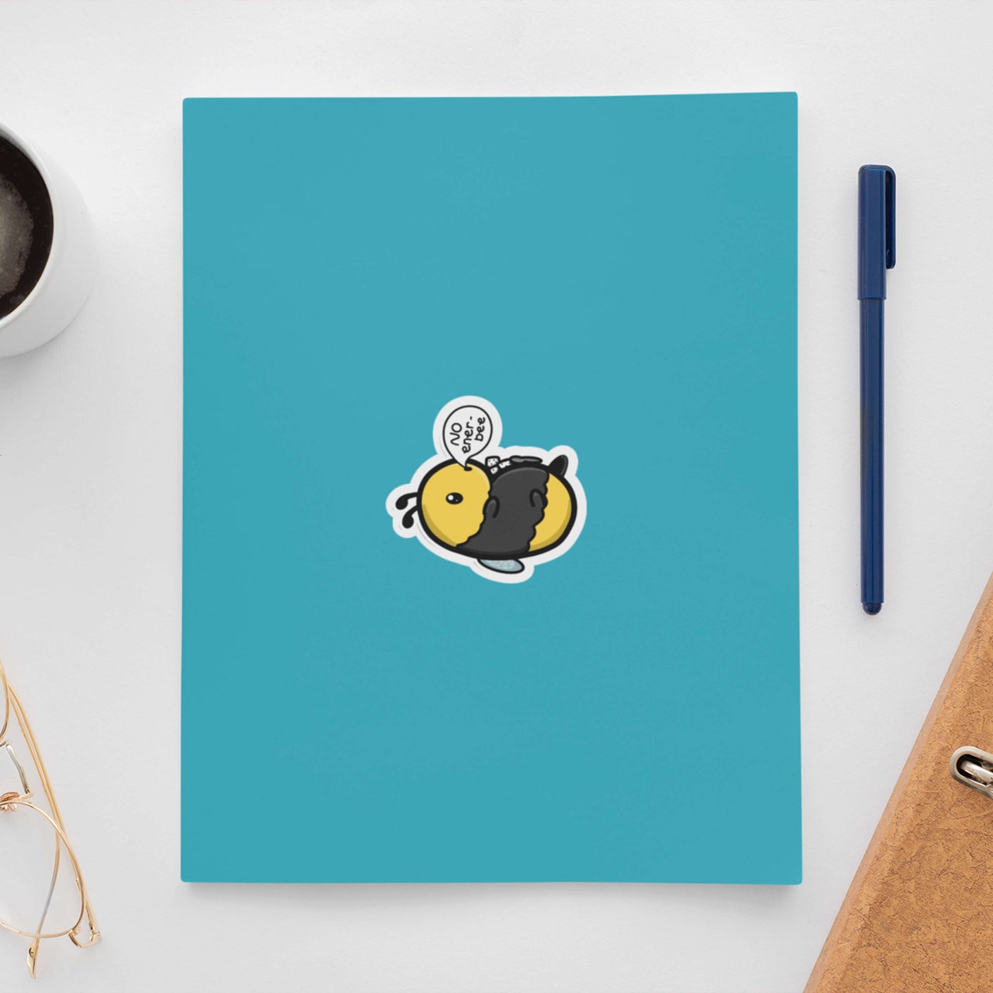 The Low Energy Bee Sticker on a blue notebook cover with a blue pen and brown folder to the right and a black coffee and gold glasses to the left. The bumble bee shaped vinyl sticker is laying on its back with sugar cubes and a spoon on its belly with a speech bubble reading 'no ener-bee'. The hand drawn design is raising awareness for chronic fatigue.