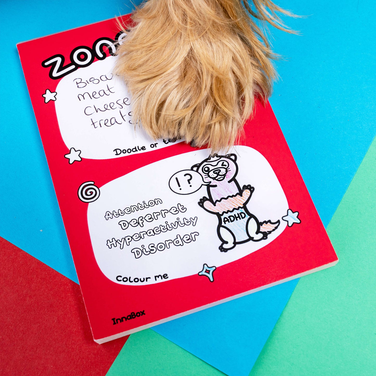 A fluffy dogs foot on top of a notepad that has an ADHD ferret on and is against a blue, green and red background