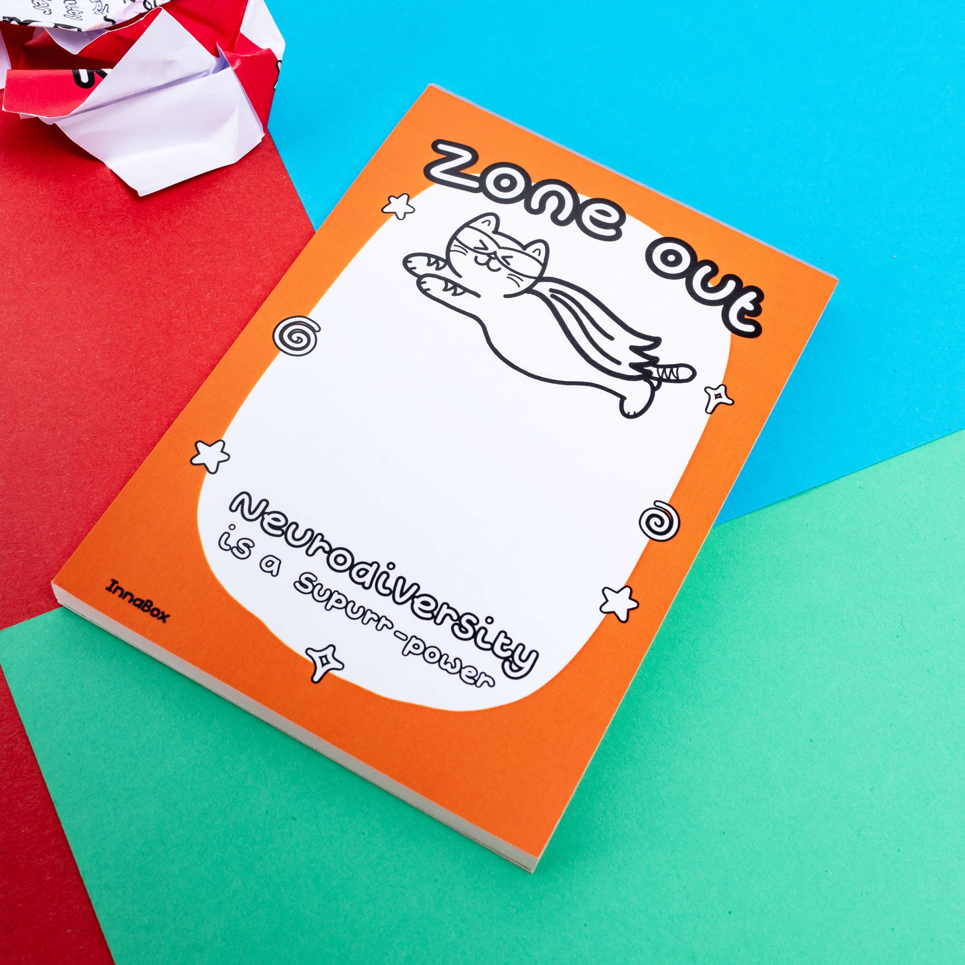 An orange page of a notepad with a superhero cat flying with the words 'Zone Out' on the top and 'Neurodiversity is a supurr-power' there is a crumpled page against a green, blue and red background.  A hand drawn design to raise awareness for neurodiversity, adhd, autism.