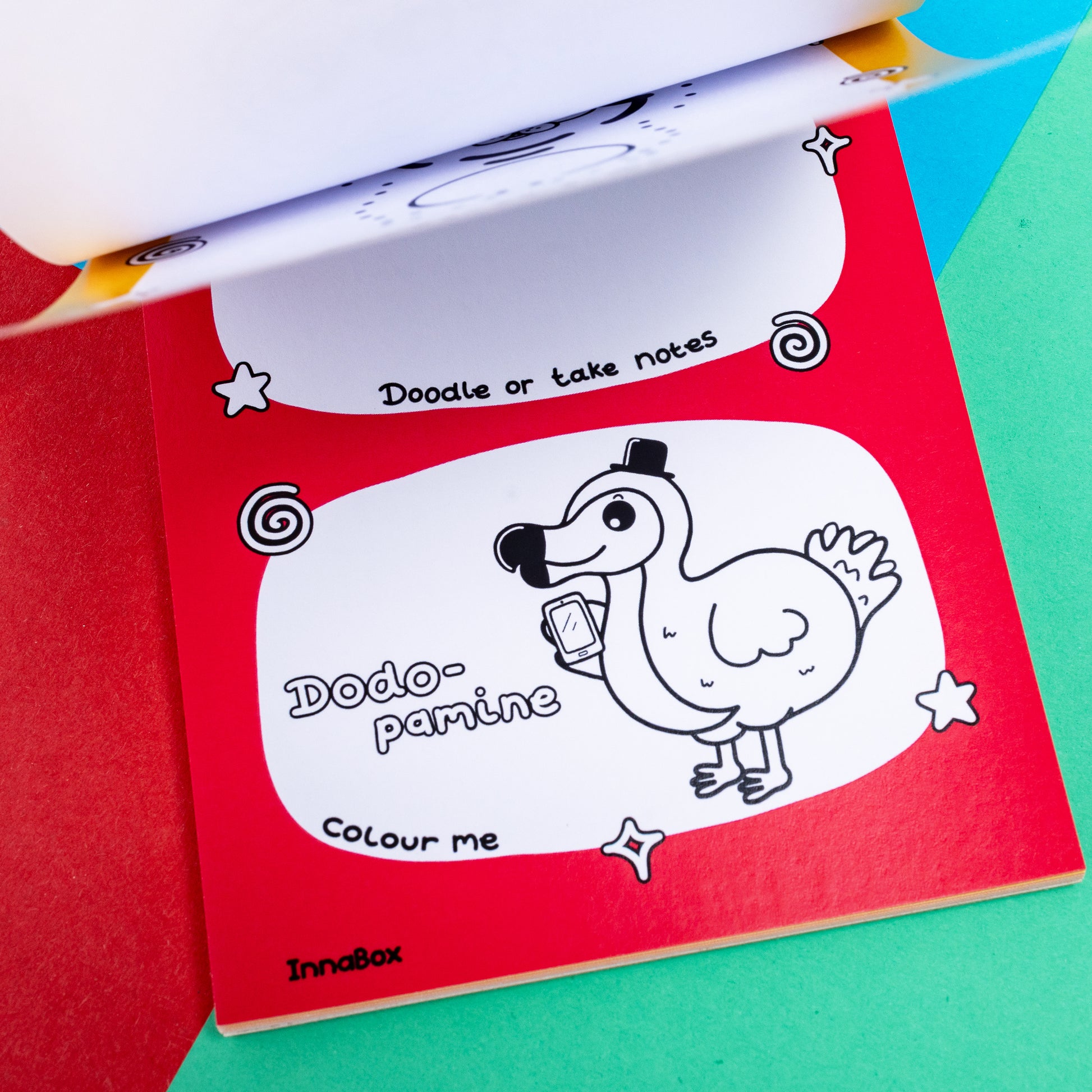 A page of a red outlined notebook with the words 'Dodo-pamine' with a Dodo drawing next to it with a phone and a top hat. The words 'Doodle or take notes' above it and below it 'colour me' it is on a red, green and blue background 