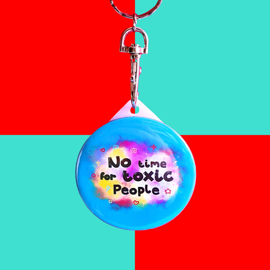 The No Time For Toxic People Keyring on a blue and red background. The silver lobster clip blue plastic circular keychain has yellow and pink clouds with black text reading 'no time for toxic people' with red, yellow, and blue hearts, clouds, sparkles, moons and flowers.