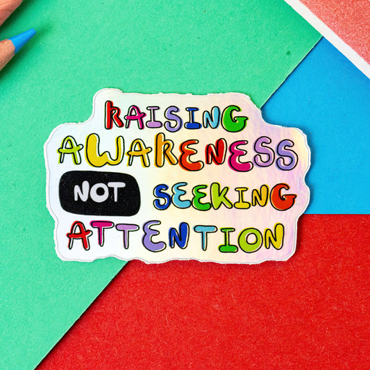 The Raising Awareness Not Seeking Attention Sticker on a red, blue and green background with colouring pencils and red stripe candy bag. The holographic sticker has rainbow bubble text reading 'raising awareness not seeking attention'.