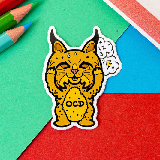 Bobsessive Compulsive Disorder Enamel Sticker - OCD on a red, green and blue background with colouring pencils and a red stripe candy bag. The bobcat shape sticker is brown with a stressed expression clutching its face with a thought bubble of numbers, question marks and lightning bolts. Across its tummy reads OCD. The sticker was designed to raise awareness of obsessive compulsive disorder.
