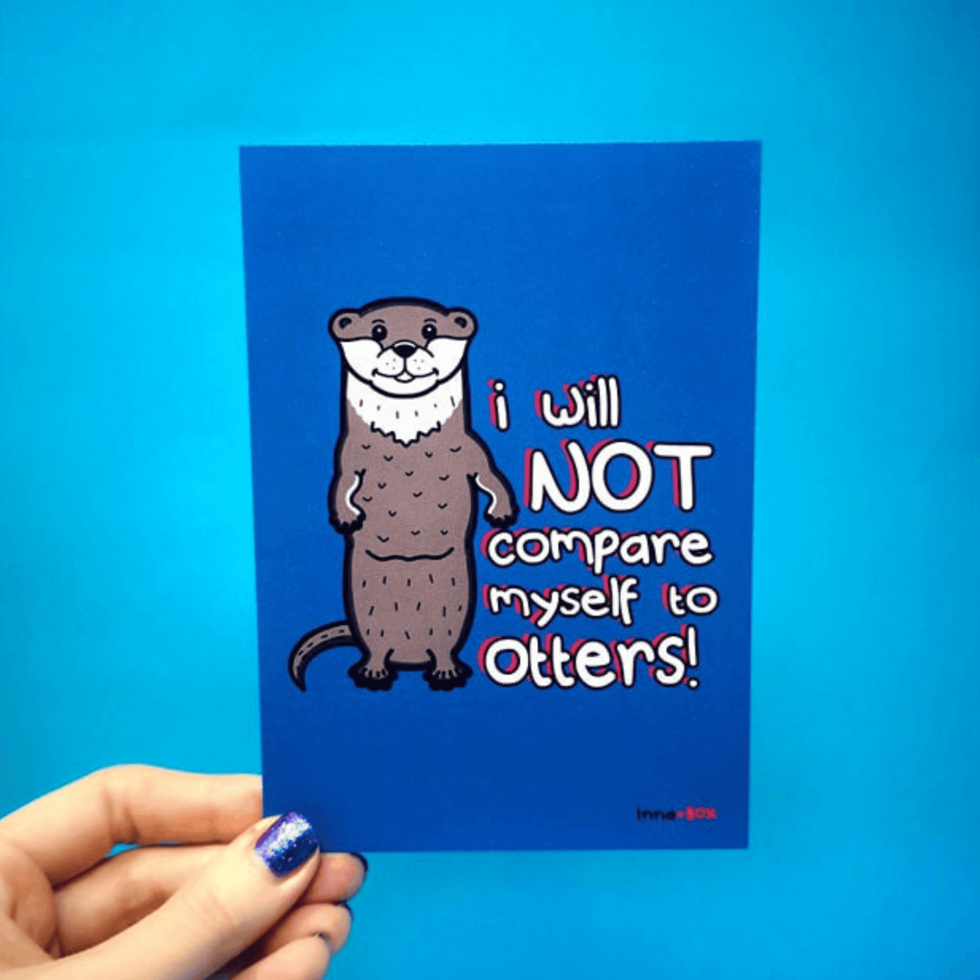 The I Will Not Compare Myself to Others Otters Postcard being held over a blue background. The blue postcard features a standing smiling grey otter with red, black and white text that reads 'I will not compare myself to otters!'. A self care and self love inspired design.