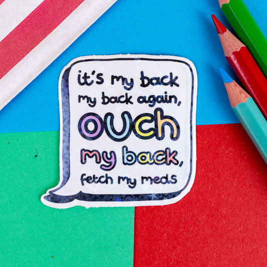 The Ouch My Back Holographic Sticker on a red, blue and green background with colouring pencils and red stripe candy bag. The holographic starry sticker is a white speech bubble with black text reading 'it's my back my back again, ouch my back, fetch my meds' with the 'ouch my back' in rainbow bubble writing.