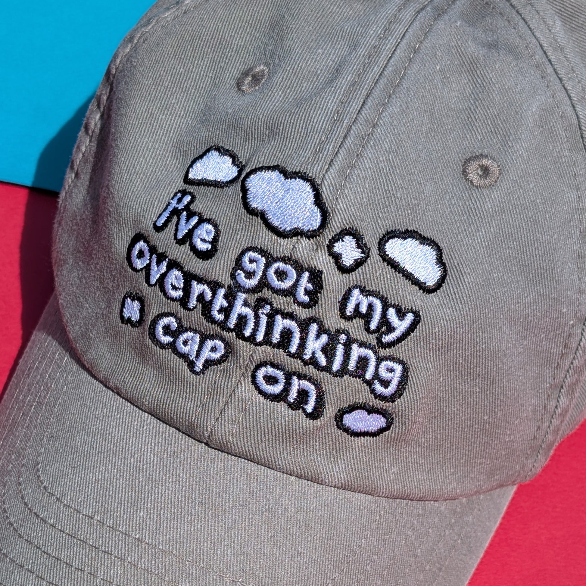 Close-up detail of the embroidery on the Innabox Overthinking Cap, laid flat on colourful blue and red paper. The text “I’ve got my overthinking cap on” is surrounded by fluffy clouds, stitched in black and white on soft grey cotton fabric.