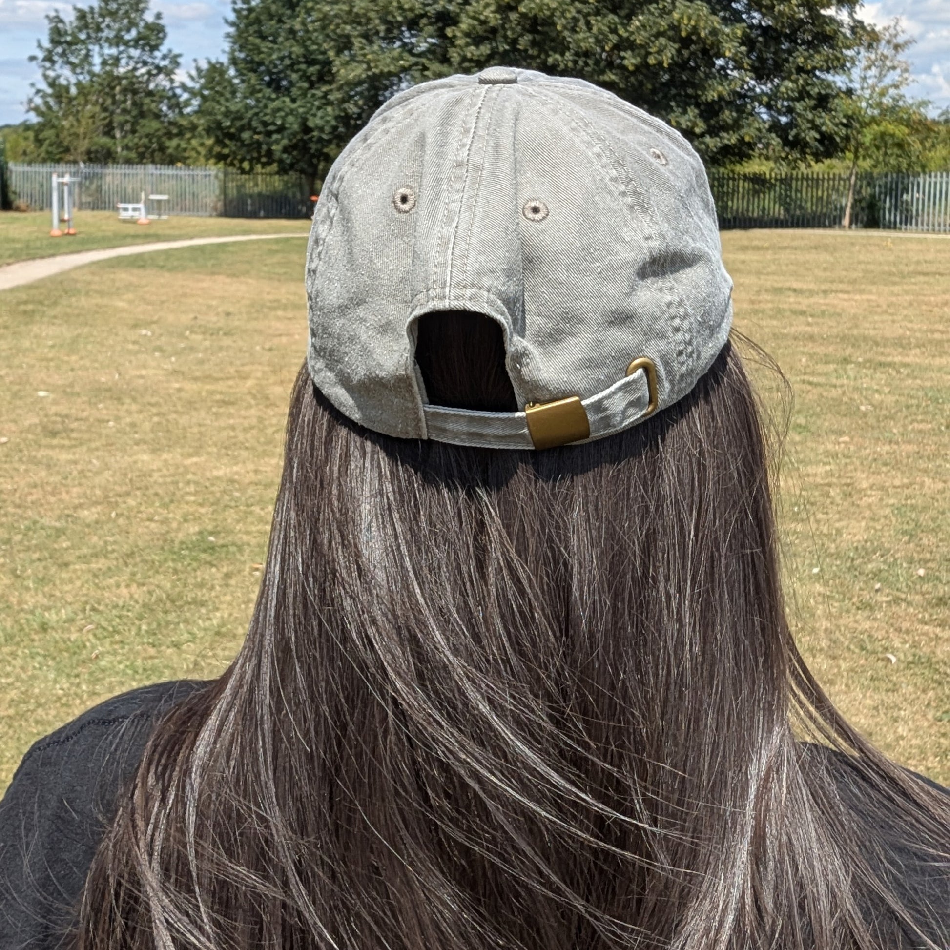 Back view of the Innabox Overthinking Cap being worn outdoors. The vintage grey cotton cap features stitched eyelets and a self-fabric strap with a brass-effect buckle for size adjustment. The person has long dark hair flowing beneath the open back.