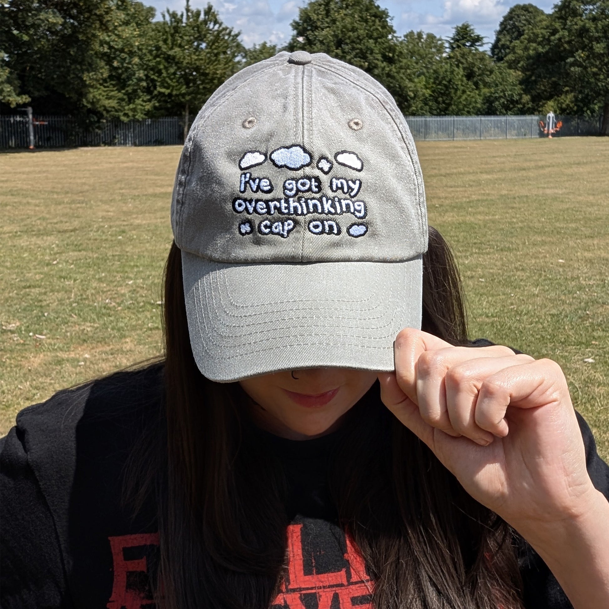 Front view of a person wearing the Overthinking Cap by Innabox, holding the peak slightly lowered. The vintage grey cap features embroidered clouds and reads “I’ve got my overthinking cap on.” Captured in a grassy park under blue skies.