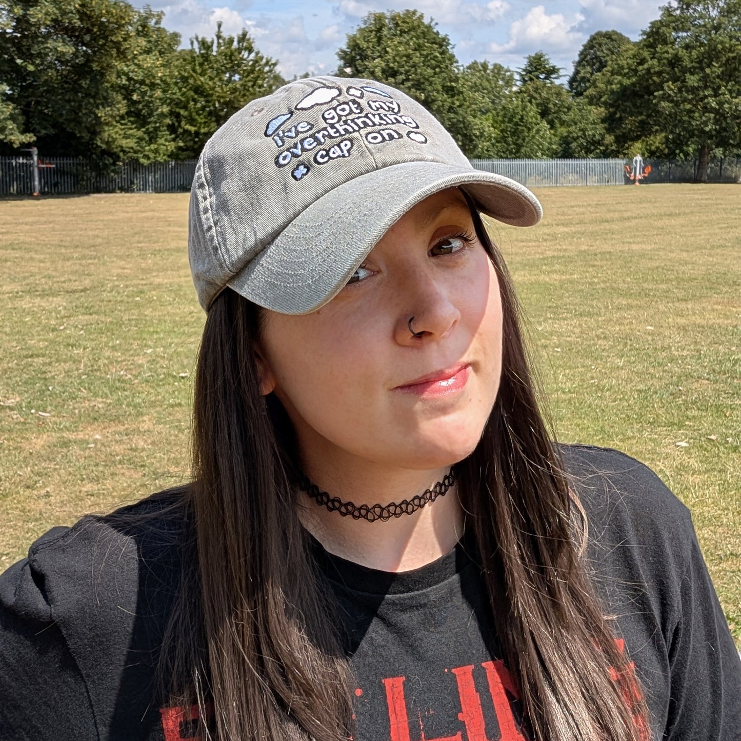 A person wearing the Innabox Overthinking Cap outdoors in a sunny park. The light grey cap reads “I’ve got my overthinking cap on” in black embroidery with small clouds. Styled with a black choker and long dark hair, the cap adds a casual, comforting vibe.