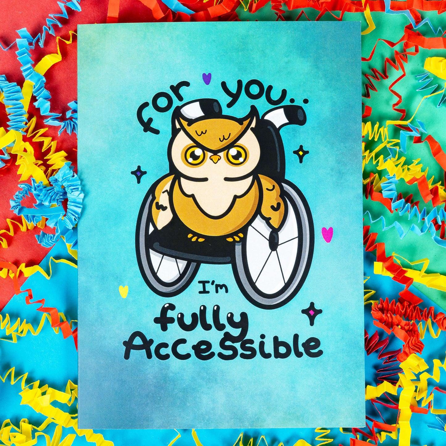 A blue greeting card with an illustration of a brown owl in a wheelchair with pink, purple and yellow love hearts and sparkles dotted around it. 'For you I'm fully accessible' is written on the card in black. The background of the photo is colourful card confetti. Design inspired by disabilities.