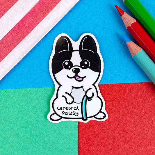 The Cerebral Pawsy Sticker - Cerebral Palsy on a red, green and blue background with colouring pencils and a red stripe candy bag. The sticker is a white and black dog with big eyes and pointy ears. The dog has it's pink tongue out and is standing on his back legs while holding a blue walking aid. 'Cerebral Pawsy' is written in black under it's tummy.