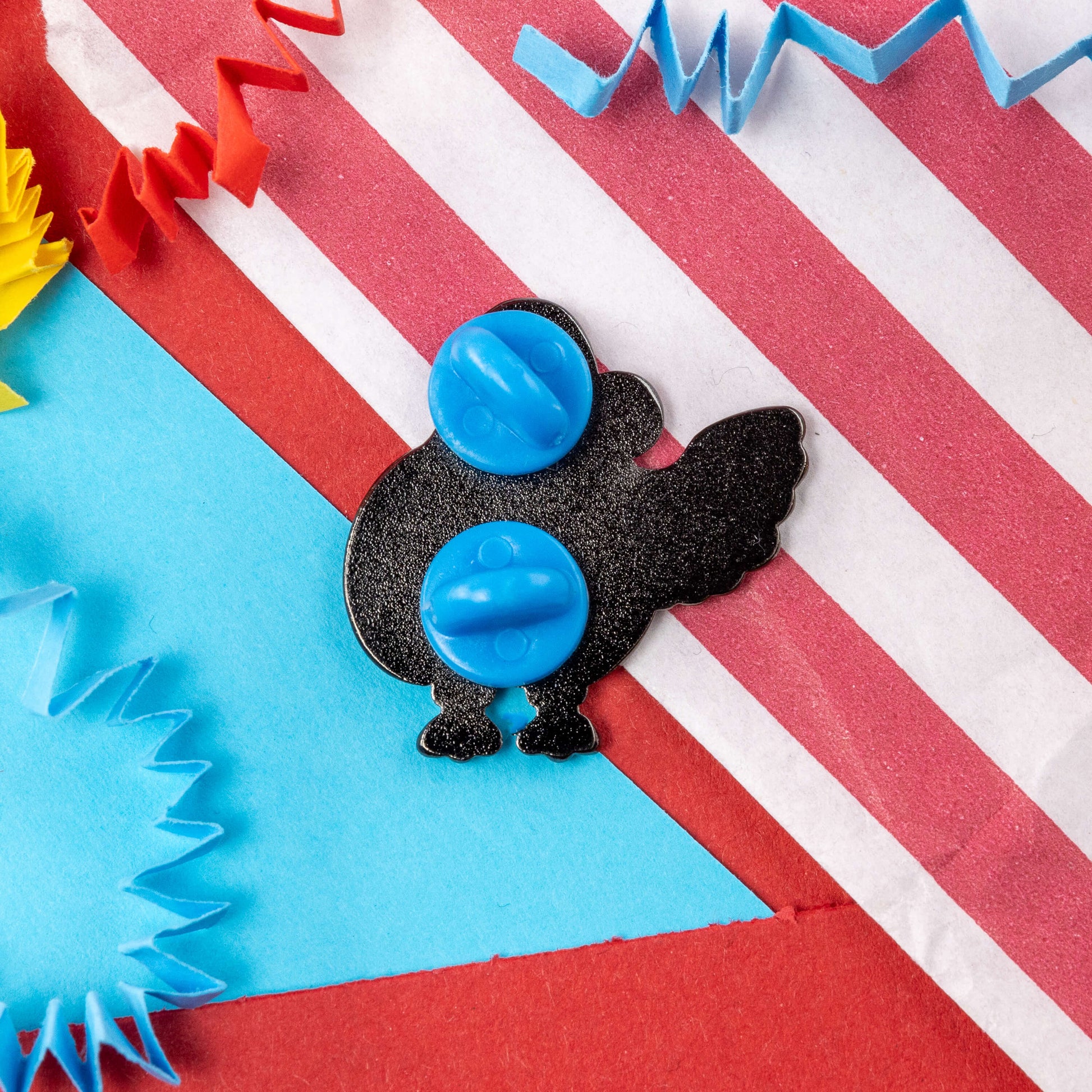 The Dyscalcoolia Pigeon Enamel Pin - Dyscalculia on a red, blue, green and yellow crinkle card confetti background. The enamel pin is facing away to show the double blue rubber backings. The enamel pin is raising awareness for dyscalculia.
