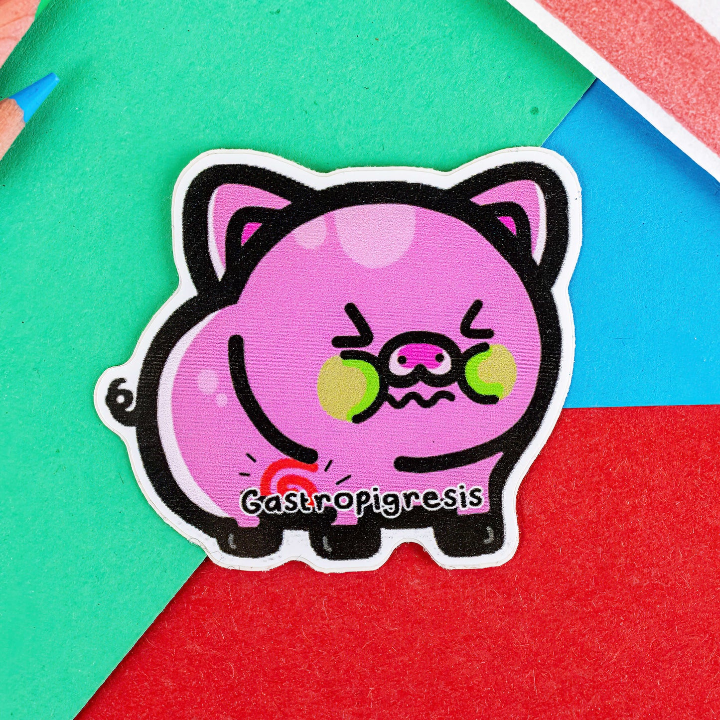 A sticker of a very pink sick looking pig with green cheeks  a swirly red tummy and Gastropigresis written across its middle. It is on a red, blue and green background with coloured pencils to one side and a stripy paper bag.
