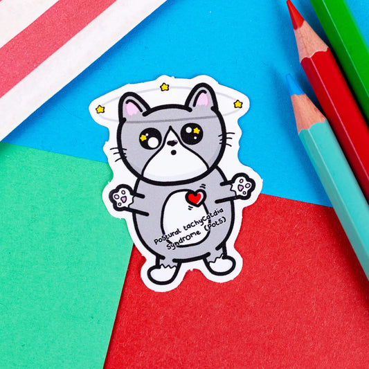 The PoTS Cat Sticker - Postural Tachycardia Syndrome on a red, blue and green background with colouring pencils and red stripe candy bag. The grey dizzy cat vinyl sticker has stars spinning above its head and in its eyes, it has a bright red heart with movement lines and black text across its middle reading 'PoStural tachyCatdid Syndrome (Pots)'.