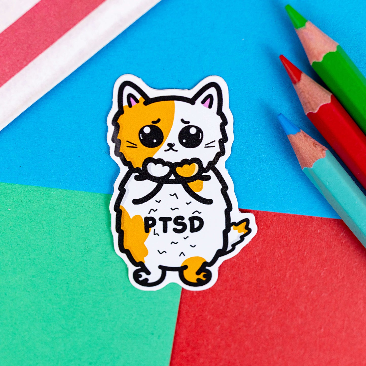 The PTSD Cat Sticker - Post-Traumatic Stress Disorder on a red, blue and green background with colouring pencils and red stripe candy bag. The orange and white scared cat sticker has black text across its middle reading 'PTSD'. The hand drawn design is raising awareness for post traumatic stress disorder.
