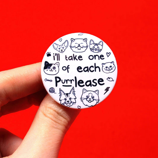 The One of Each Cat Purrlease Badge/Magnet being held over a red background. The white circular button badge or magnet has 'I'll take one of each purrlease' in black in the middle with outlines of different breeds of cat faces smiling, cat toys, hearts and paw prints.