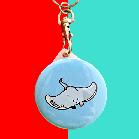 The Raynaud's Sting Ray Keyring on a red and blue background. The silver lobster clip blue plastic circular keychain with a grey sting ray with a scrunched sad face and red patches with black text across its wing reading 'raynaud's'. The hand drawn design is raising awareness for Raynaud's.