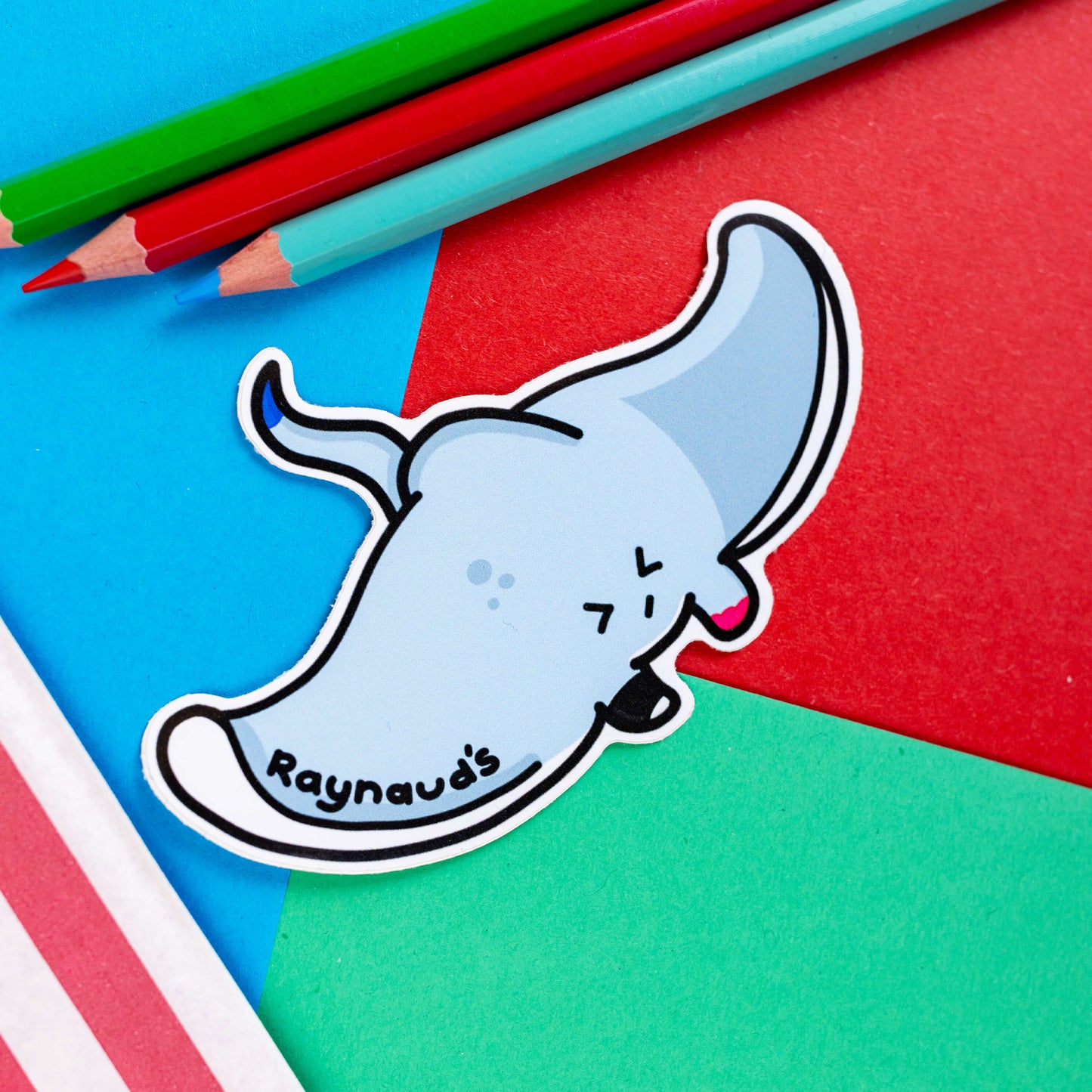 The Raynaud's Sting Ray Sticker on a red, blue and green background with colouring pencils and red stripe candy bag. The pastel blue grey stingray sticker has a scrunched sad face with red patches and black text across its wing reading 'raynaud's'. The hand drawn design is raising awareness for Raynaud's disease.
