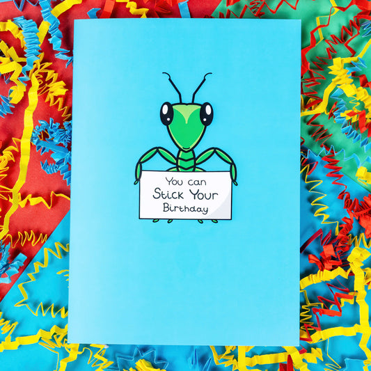 The You Can Stick Your Birthday Insect Card on a blue, green and red background with blue, yellow and red crinkle card confetti. The sky blue card has a green stick insect in the centre of it with big shiny eyes and two antennae on its head. The stick insect is holding a white sign that reads 'You can Stick Your Birthday' in black writing.
