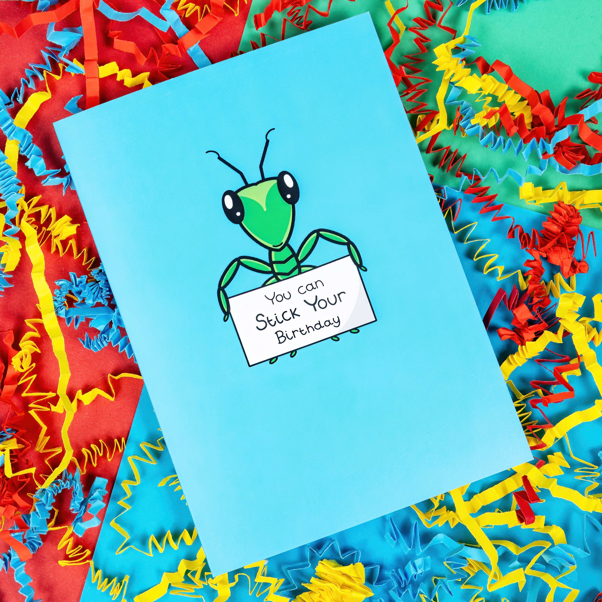 The You Can Stick Your Birthday Insect Card on a blue, green and red background with blue, yellow and red crinkle card confetti. The sky blue card has a green stick insect in the centre of it with big shiny eyes and two antennae on its head. The stick insect is holding a white sign that reads 'You can Stick Your Birthday' in black writing.