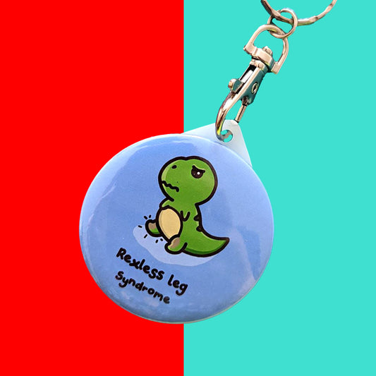 The Rexless Leg Syndrome Keyring - Restless Leg Syndrome on a red and blue background. The silver lobster clip plastic blue circular keychain features an angry upset green t-rex dinosaur with red lower legs and black lines, underneath in black text reads 'rexless leg syndrome'. The hand drawn design is raising awareness for restless legs syndrome.