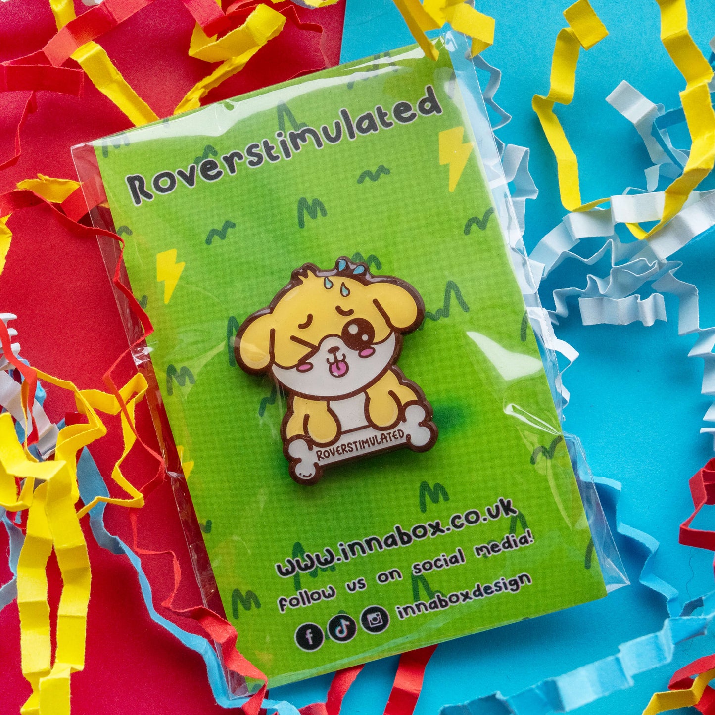 Roverstimulated Dog enamel pin by Innabox displayed on bright green backing card with colourful red, yellow and blue paper streamers. 30mm hard enamel pin featuring a cute yellow dog holding a bone that reads “Roverstimulated”, representing overstimulation and hidden disabilities awareness.
