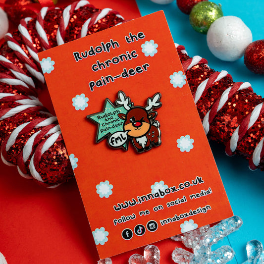 Innabox pin of 'Rudolph the Chronic Pain-deer' on a red card surrounded by festive decorations, offering a light-hearted approach to chronic pain awareness and hidden disability support.