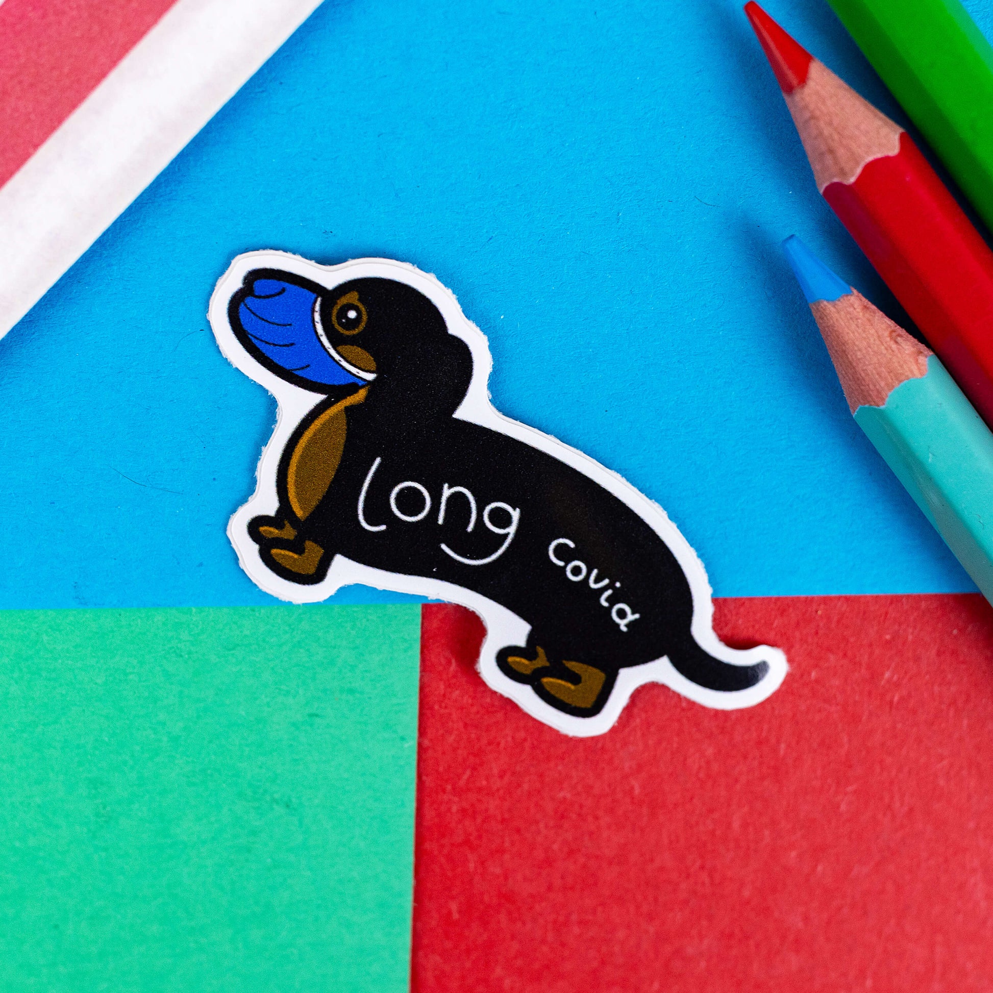 A brown and black sausage dog sticker wearing a blue medical mask over mouth and nose and 'long covid' written across it's body in white. The sticker is shown on a red and blue background. The hand drawn design is raising awareness for long covid corona virus.