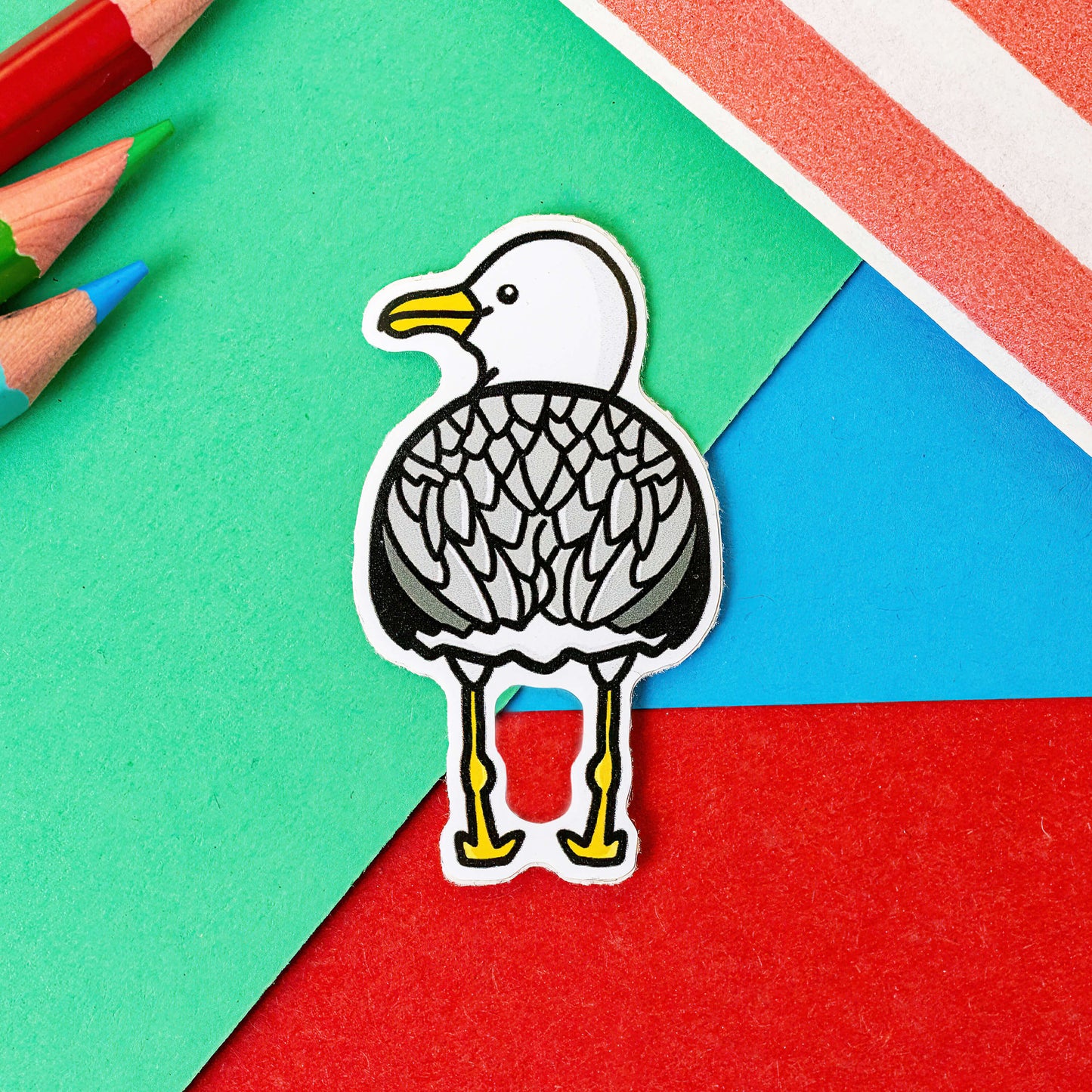 The Seagull Bum Sticker on a red, blue and green background with colouring pencils and red stripe candy bag. The seagull shape vinyl sticker is facing away showing off its bum smiling looking over its shoulder. 