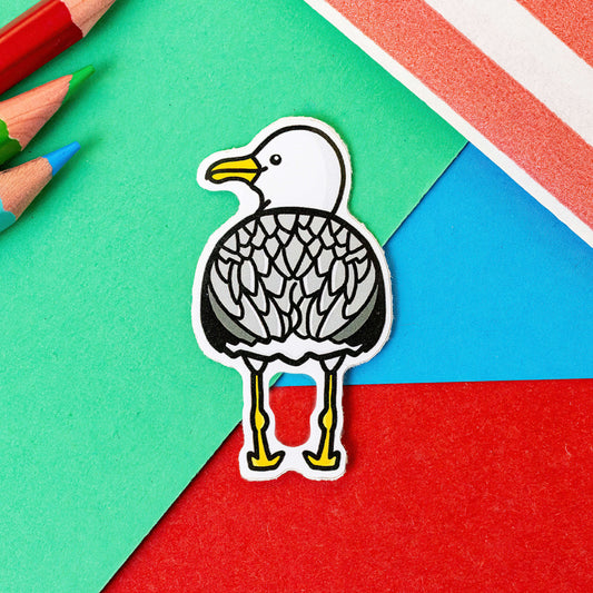 The Seagull Bum Sticker on a red, blue and green background with colouring pencils and red stripe candy bag. The seagull shape vinyl sticker is facing away showing off its bum smiling looking over its shoulder. 