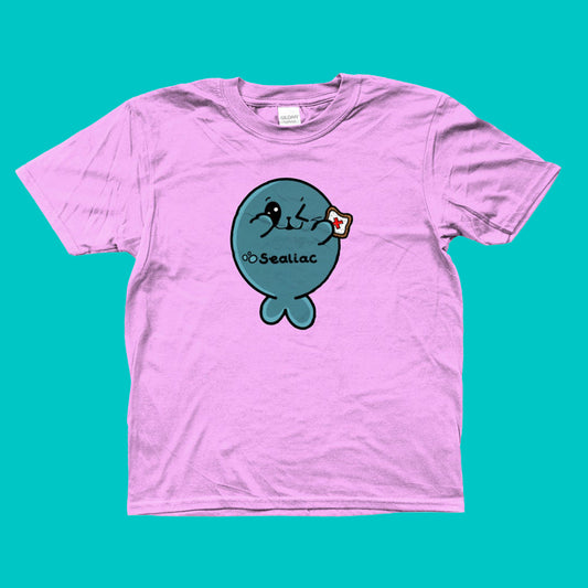 The Sealiac Coeliac Kids Tee - Coeliac on a red and blue background. The light pink short sleeve tshirt features a winking blue seal holding up a piece of toast with a red cross in the middle, across its belly in black reads 'Sealiac'. The hand drawn design is raising awareness for Coeliac and gluten allergy.