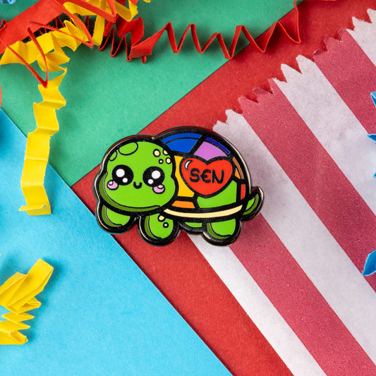 The Speshell Educational Needs Enamel Pin - SEN - Special Educational Needs on a red, blue, yellow and green card confetti background. A rainbow shell kawaii cute style tortoise with pink cheeks and sparkling eyes, on its shell is a red heart with 'SEN' in the middle. The pin design is raising awareness for SEN Special Educational Needs.