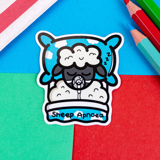 The Sheep Apnoea Sticker - Sleep Apnoea on a red, blue and green background with colouring pencils and a red stripe candy bag. The sticker features a sleeping sheep wearing a CPAP mask laying on a blue cloud pillow and duvet, across the duvet in black reads 'sheep apnoea'. The hand drawn design is raising awareness for Sleep apnoea.