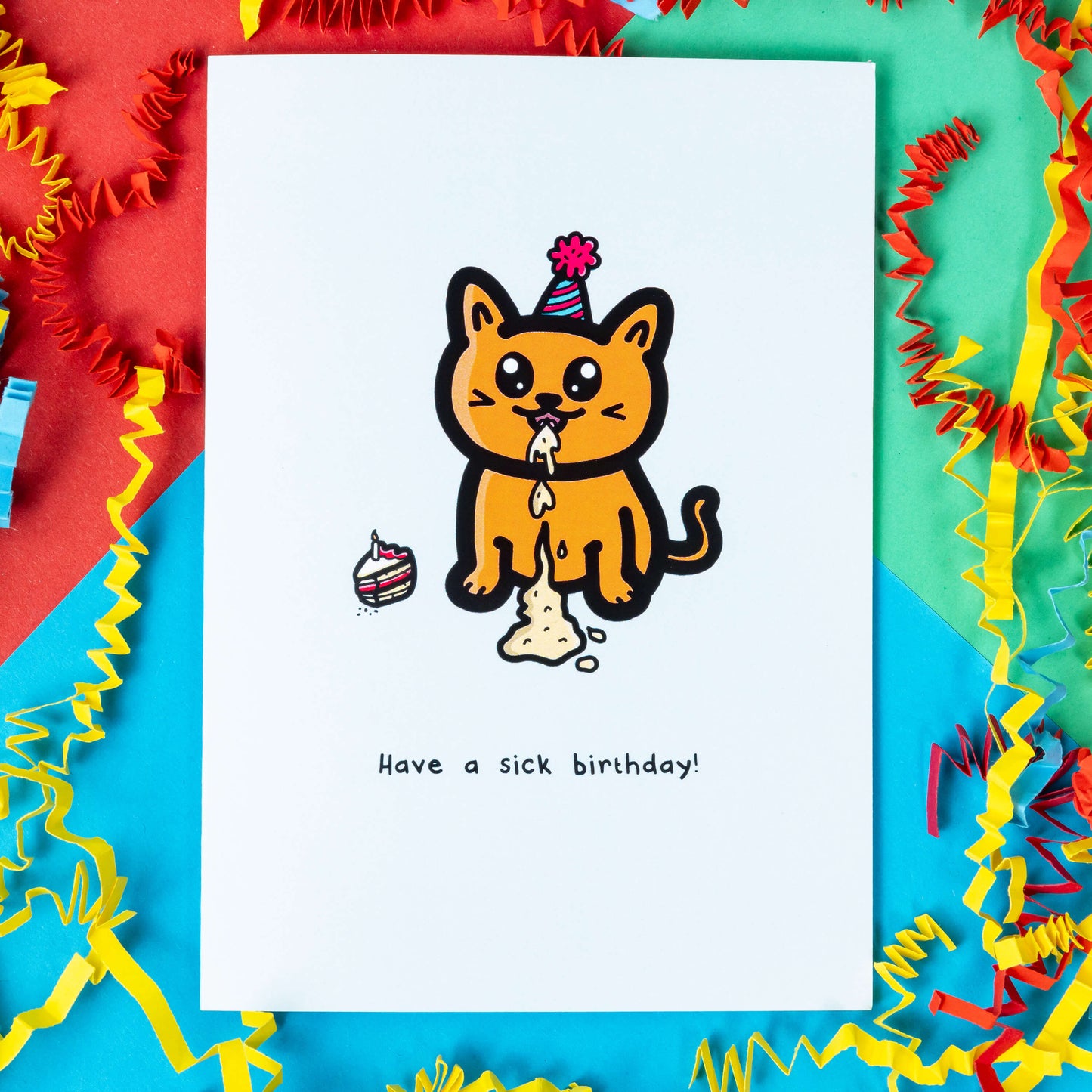 The Have a Sick Birthday Cat Card on a red, blue and green background with red, yellow and blue crinkle card confetti. The white a6 birthday card has an orange smiling cat with a party hat throwing up vomit all down itself next to a birthday cake slice, underneath in black reads 'have a sick birthday!'. 