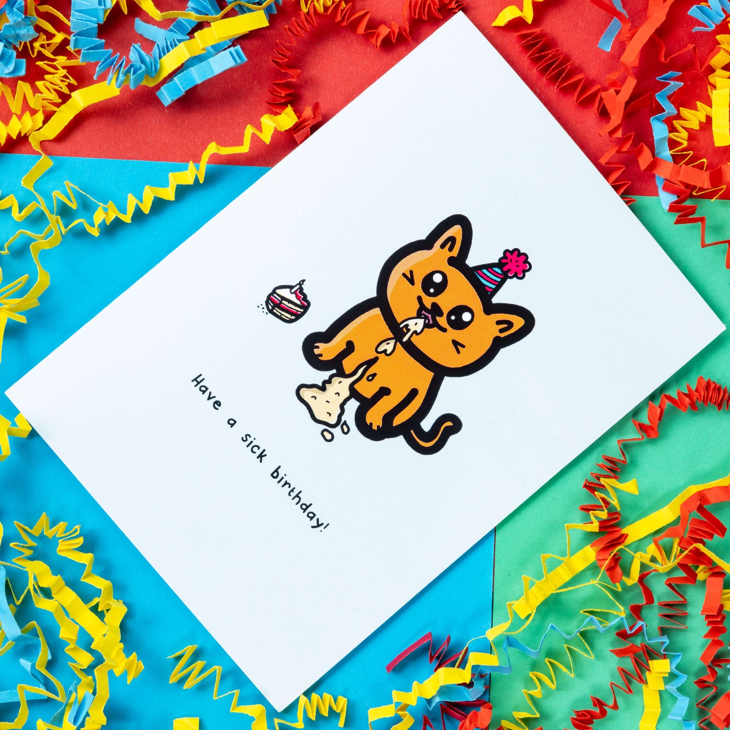 The Have a Sick Birthday Cat Card on a red, blue and green background with red, yellow and blue crinkle card confetti. The white a6 birthday card has an orange smiling cat with a party hat throwing up vomit all down itself next to a birthday cake slice, underneath in black reads 'have a sick birthday!'.