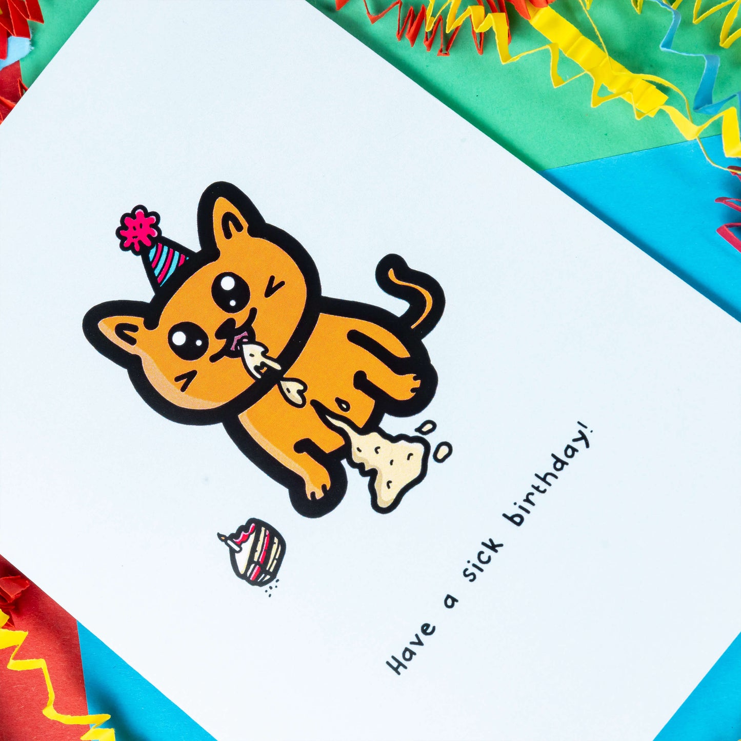 The Have a Sick Birthday Cat Card on a red, blue and green background with red, yellow and blue crinkle card confetti. The white a6 birthday card has an orange smiling cat with a party hat throwing up vomit all down itself next to a birthday cake slice, underneath in black reads 'have a sick birthday!'.