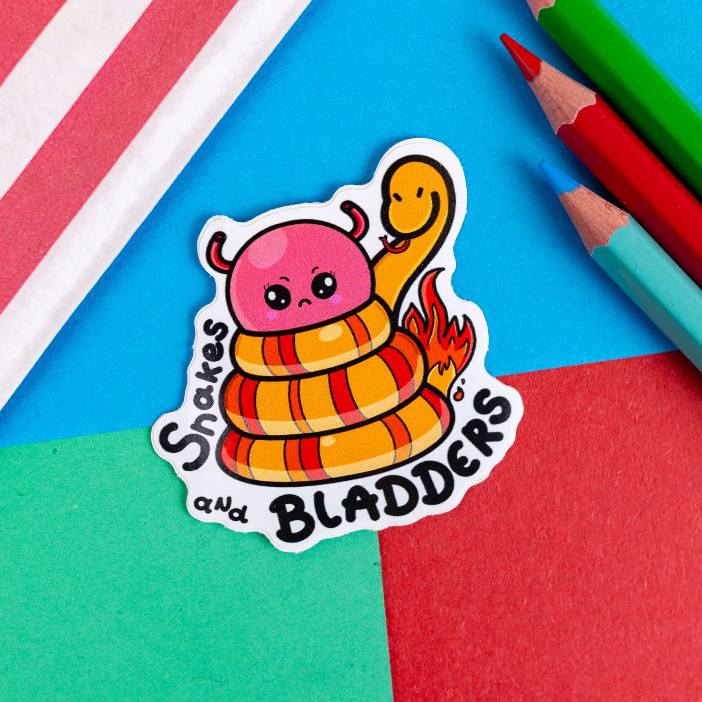A sticker with a snake wrapped around an angry looking bladder with a face with the words snakes and bladders around the bottom. The sticker is laid on a blue, green and red background with pencils and a striped bag in the corners