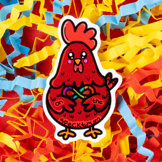 A red cartoon chicken sticker with a rainbow infinity symbol on its belly, holding the symbol with its wings. The text "on the specktrum" is written on the chicken's body. The sticker is placed on a background of colorful shredded paper in yellow, red, and blue. Hand drawn design created to raise awareness for ASD and neurodivergence