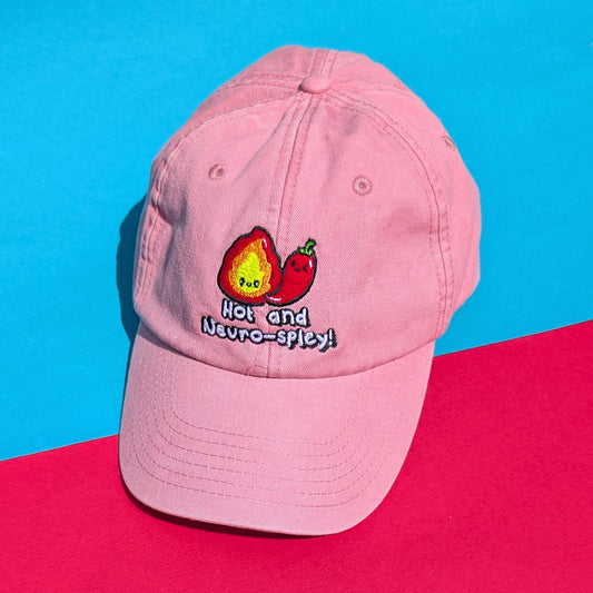 Pink cotton cap with embroidered flame and chilli design reading "Hot and Neuro-Spicy!" by Innabox, laid flat on bright red and blue background.